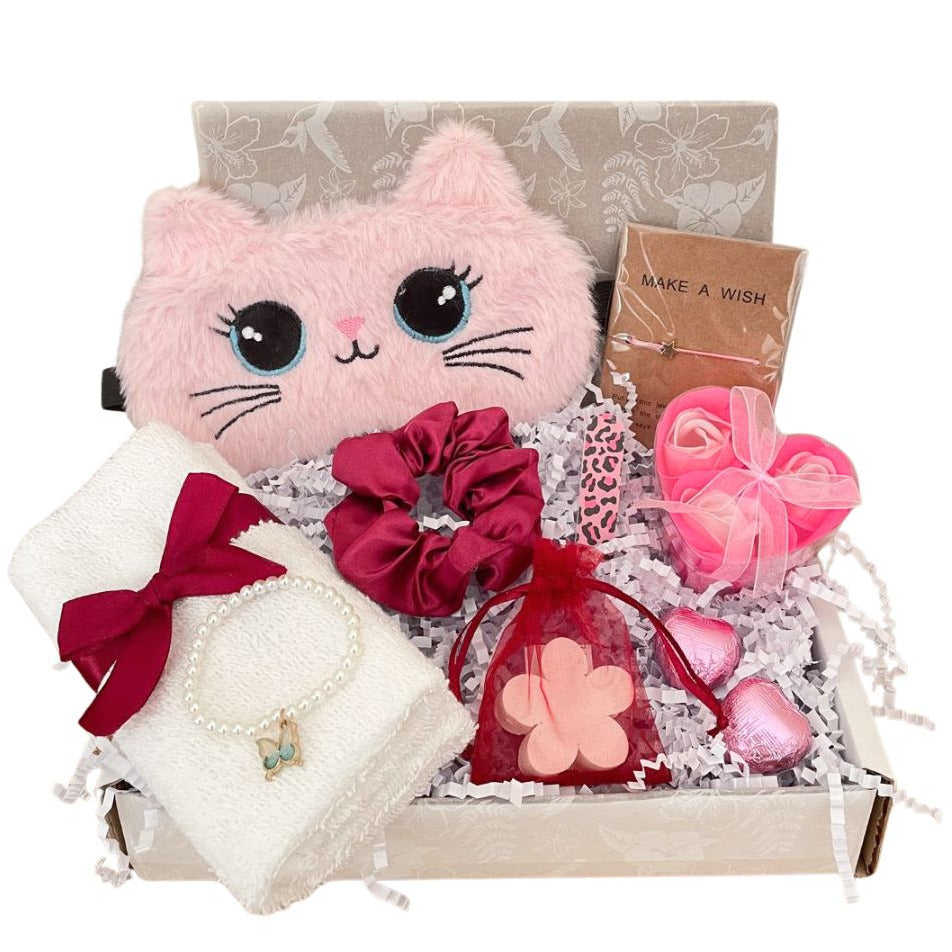 A teenage girls pamper gift box containing a pink plush cat sleep mask, a white towel with a dark pink ribbon, a dark pink satin scrunchie, a bottle of Graceful Orchid fragrance mist, two heart-shaped sweets, and a Make A Wish bracelet card.