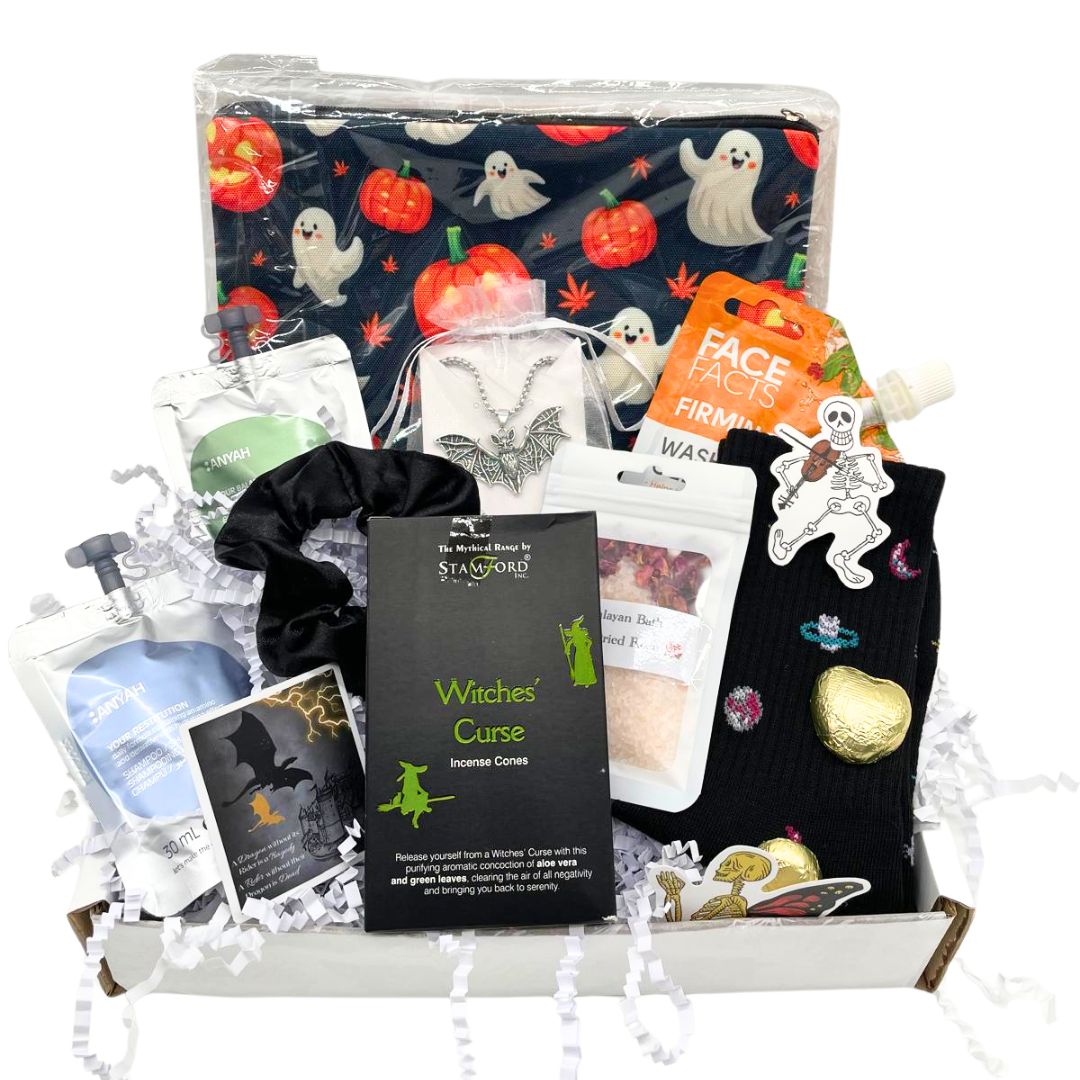A Halloween-themed gift box filled with assorted witch and spooky treats. The box contains a black witch-themed pouch with a ghost and pumpkin pattern, a black scrunchie, black socks, silver bat charm necklace, a card, shampoo, body wash, chocolates all arranged on white crinkle paper inside a white box.