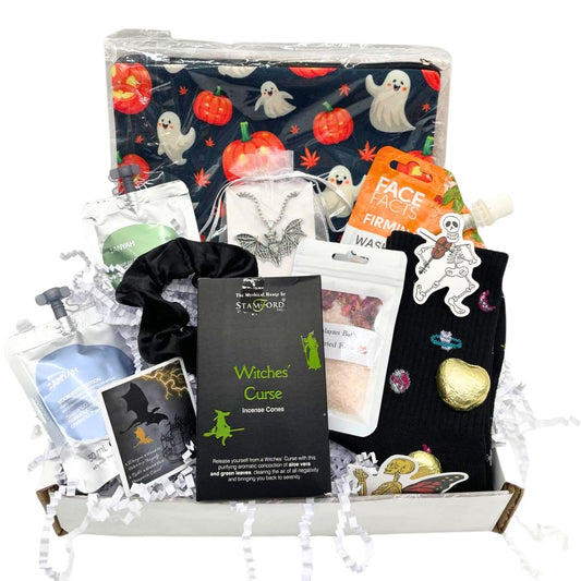 A Halloween-themed gift box filled with assorted witch and spooky treats. The box contains a black witch-themed pouch with a ghost and pumpkin pattern, a black scrunchie, black socks, silver bat charm necklace, a card, shampoo, body wash, chocolates all arranged on white crinkle paper inside a white box.