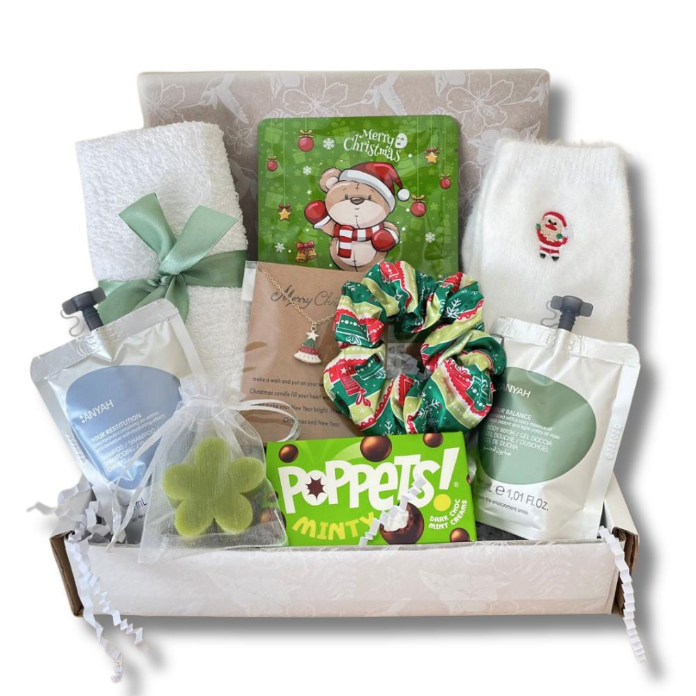 A curated Teenage gift box filled with teen care items including a green Christmas-themed socks, a mint green scrunchie, a green clover-shaped bath soa, and two white pouches of bath products, all arranged in a white tray with shredded paper.