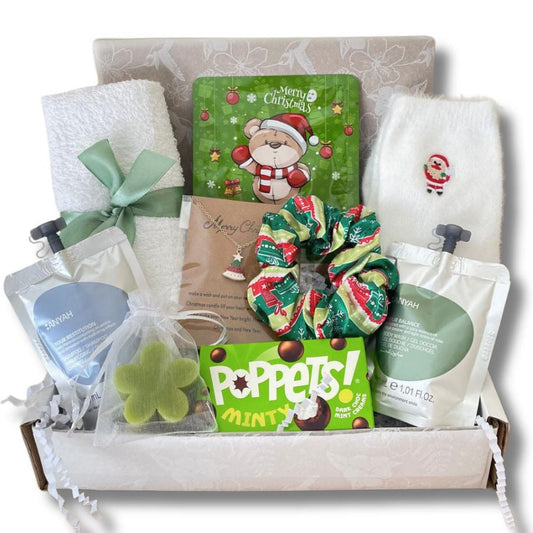 A curated Teenage gift box filled with teen care items including a green Christmas-themed socks, a mint green scrunchie, a green clover-shaped bath soa, and two white pouches of bath products, all arranged in a white tray with shredded paper.