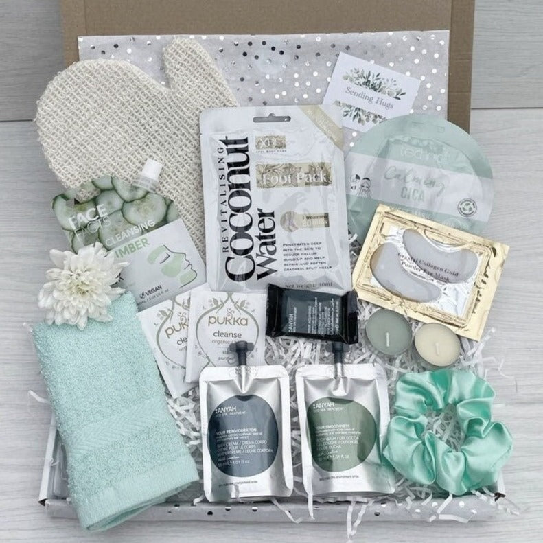 A gift box containing various beauty products including a green and white spa set, a coconut foot pack, a face mask, a scrub mitten, a satin scrunchie, a gift box  and card.