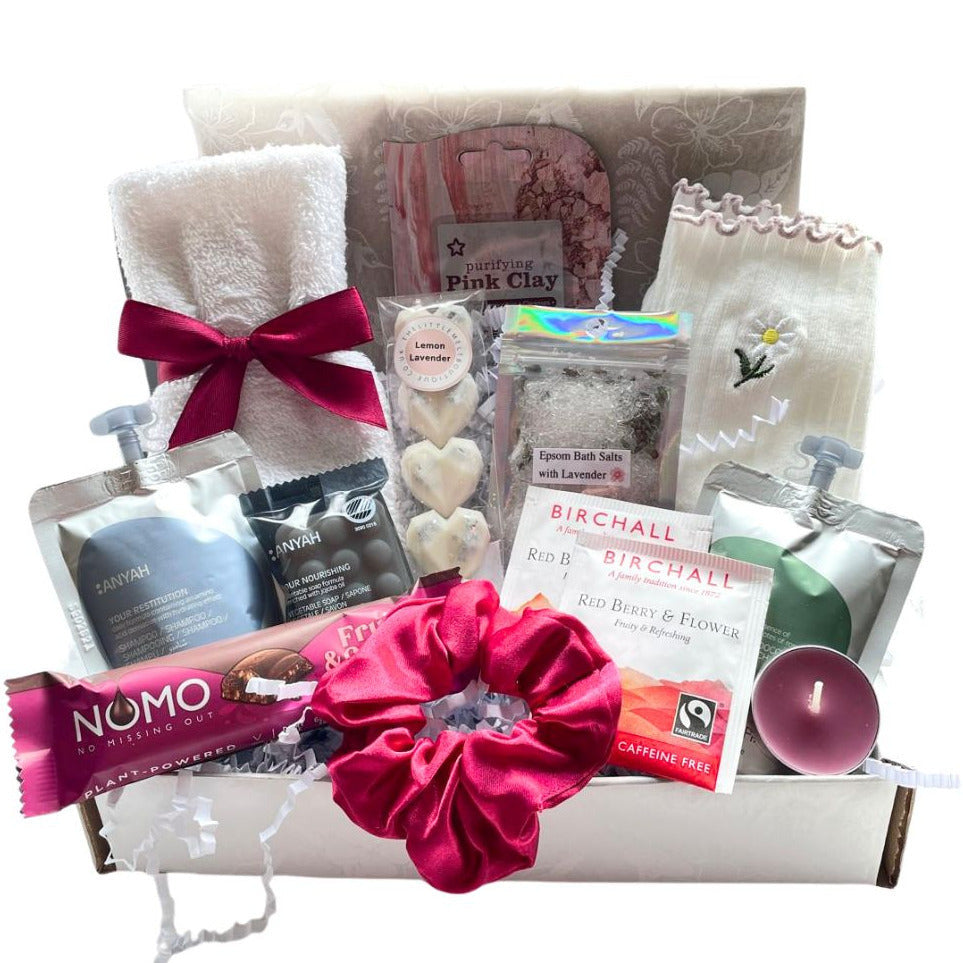 A gift box containing a white towel with a burgundy ribbon, a pink clay face mask, a burgundy scrunchie, a card reading Just For You, two Birchall tea bags, a Nomo chocolate bar, a bag of Epsom salts, and white socks with a daisy.