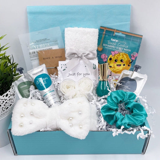 A gift box containing various spa-related items such as a face mask, nail polish, body wash, body lotion, rose flower soaps, a bracelet, hair accessories, a face towel, and hand cream, all packaged in a decorative teal box.
