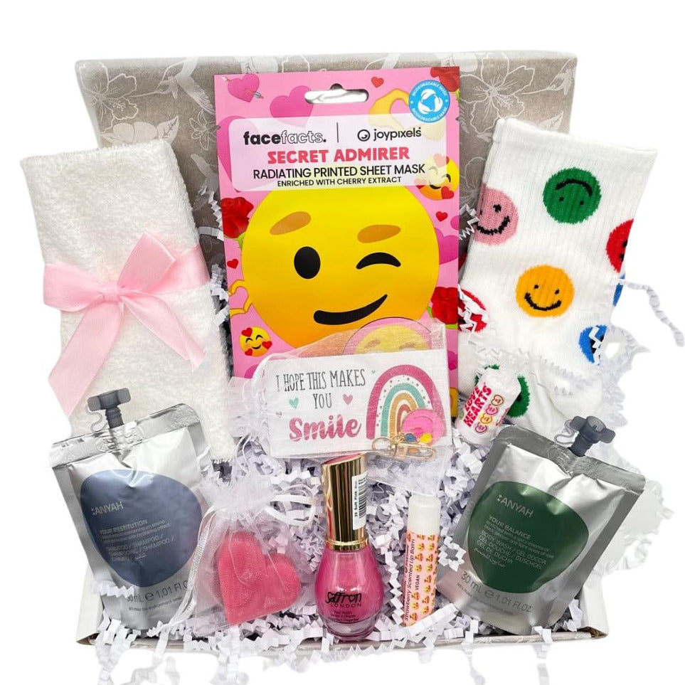 A curated gift set in a cardboard tray filled with white crinkle paper, featuring a pink bow on a white towel, a winking emoji face mask pack, a small pink nail polish bottle, a lip balm, and two branded wash and shampoo  in foil pouches.