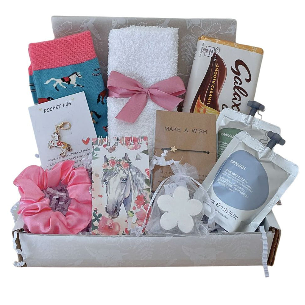 A curated gift box filled with assorted self-care and beauty items, including a pink scrunchie, a white towel tied with a pink bow, horse patterned socks, a notebook with a horse illustration, a small soap, a pouch with shampoo, and other gift items, all arranged in a light gray box on a white background.