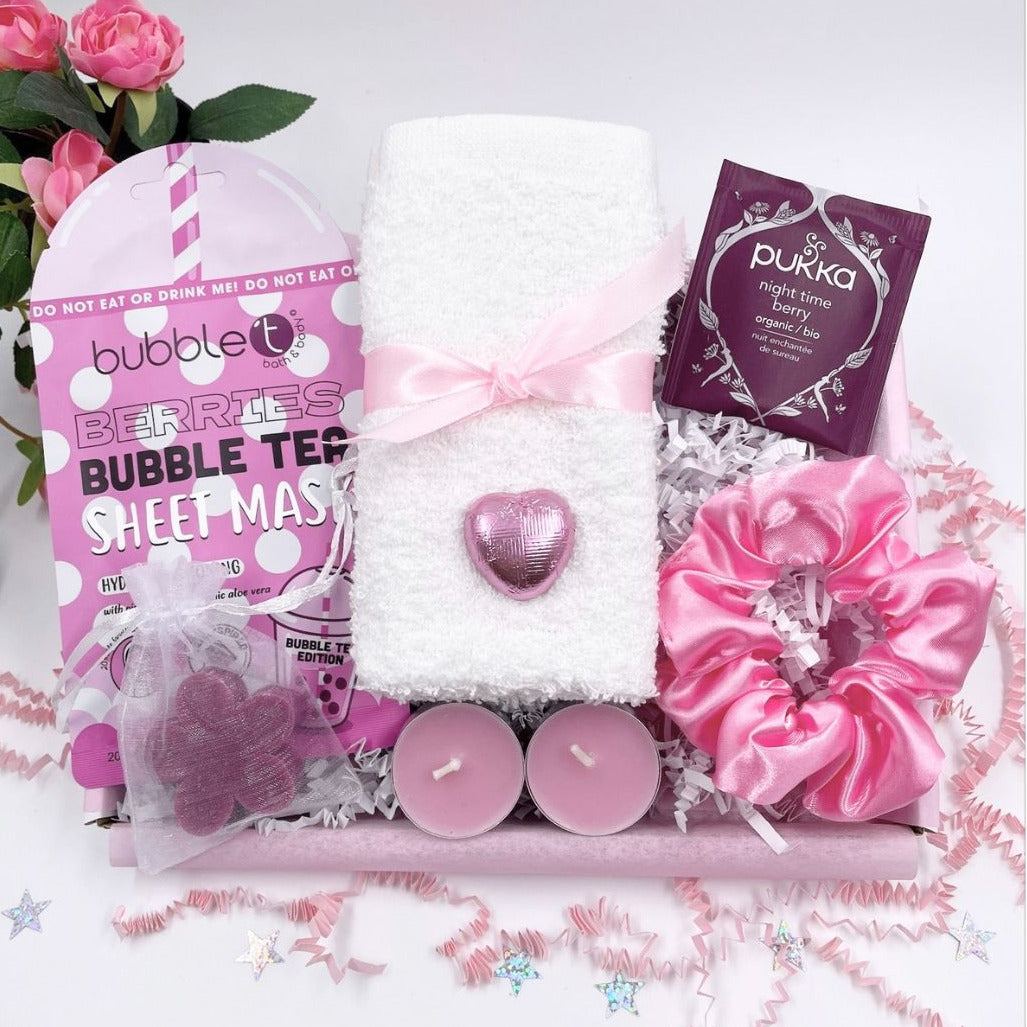A pink-themed self-care gift set arranged in a tray with a white towel tied with a pink ribbon, a pink heart-shaped soap, two pink candles, a pink satin scrunchie, a bubble tea packet, and a sheet mask package, with pink roses and confetti around it.