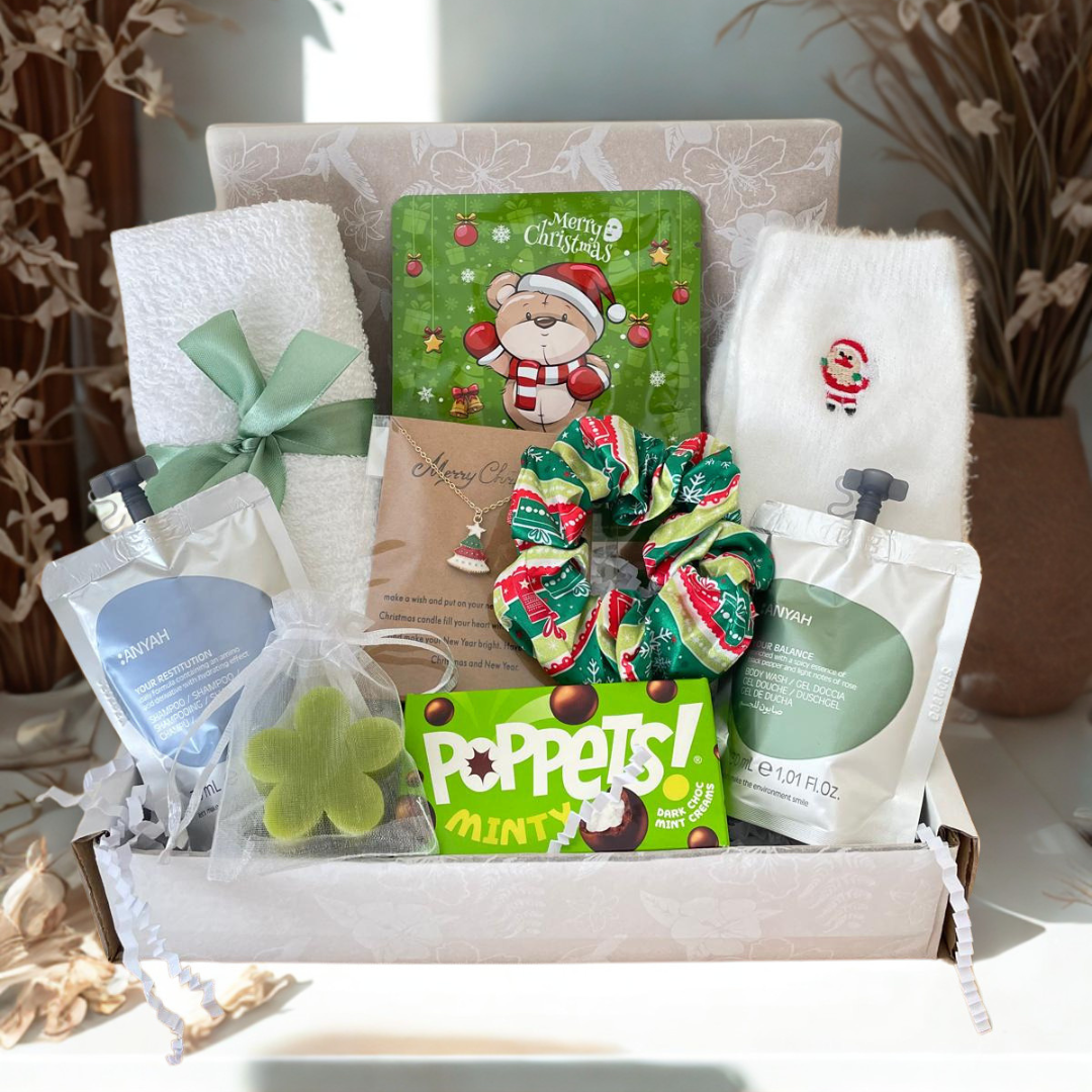 A curated gift box containing a white towel with a mint green ribbon, a green Christmas-themed tin, a patterned green and red scrunchie, a bag of bath salts, a necklace, and a box of Poppets! Minty Snacks, arranged in white tissue paper.