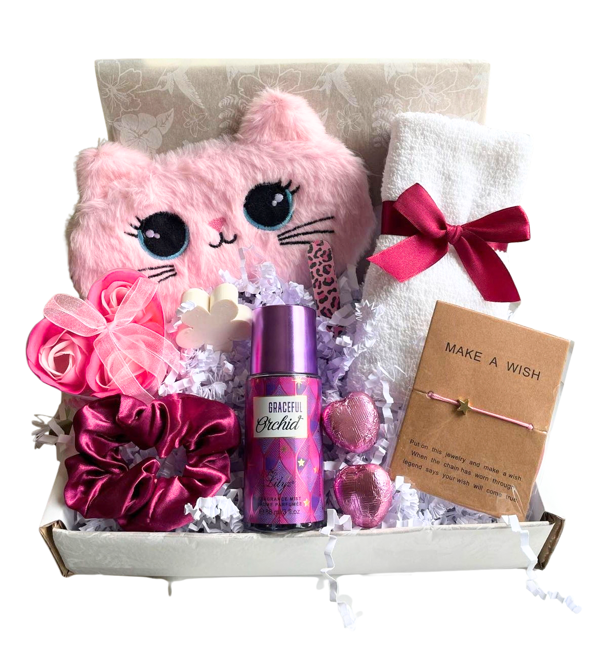 A teenage girls pamper gift box containing a pink plush cat sleep mask, a white towel with a dark pink ribbon, a dark pink satin scrunchie, a bottle of Graceful Orchid fragrance mist, two heart-shaped sweets, and a Make A Wish bracelet card.
